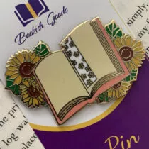 detailfoto van pin, opengeslagen boek met aan de linker en rechterkant 2 zonnebloemen en in het midden een boekenlegger met kleine gele bloemen