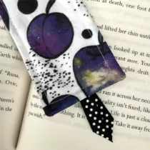 detailfoto van witte boekenlegger met appels met galaxy kleuren, paars, geel, blauw. Met zwart lint met witte stippen aan de onderkant