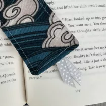 detailfoto van boekenlegger met donkerblauwe achtergrond en witte wolken. Grijs lintje met witten stippen aan de onderkant