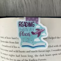 detailfoto van de magnetische boekenlegger met de tekst reading takes you places, op een blauwe achtergrond met een open boek en vliegtuig.
